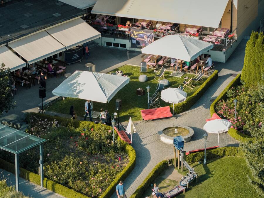 An outdoor dining area with tables and umbrellas, surrounded by a garden with hedges and flowers.
