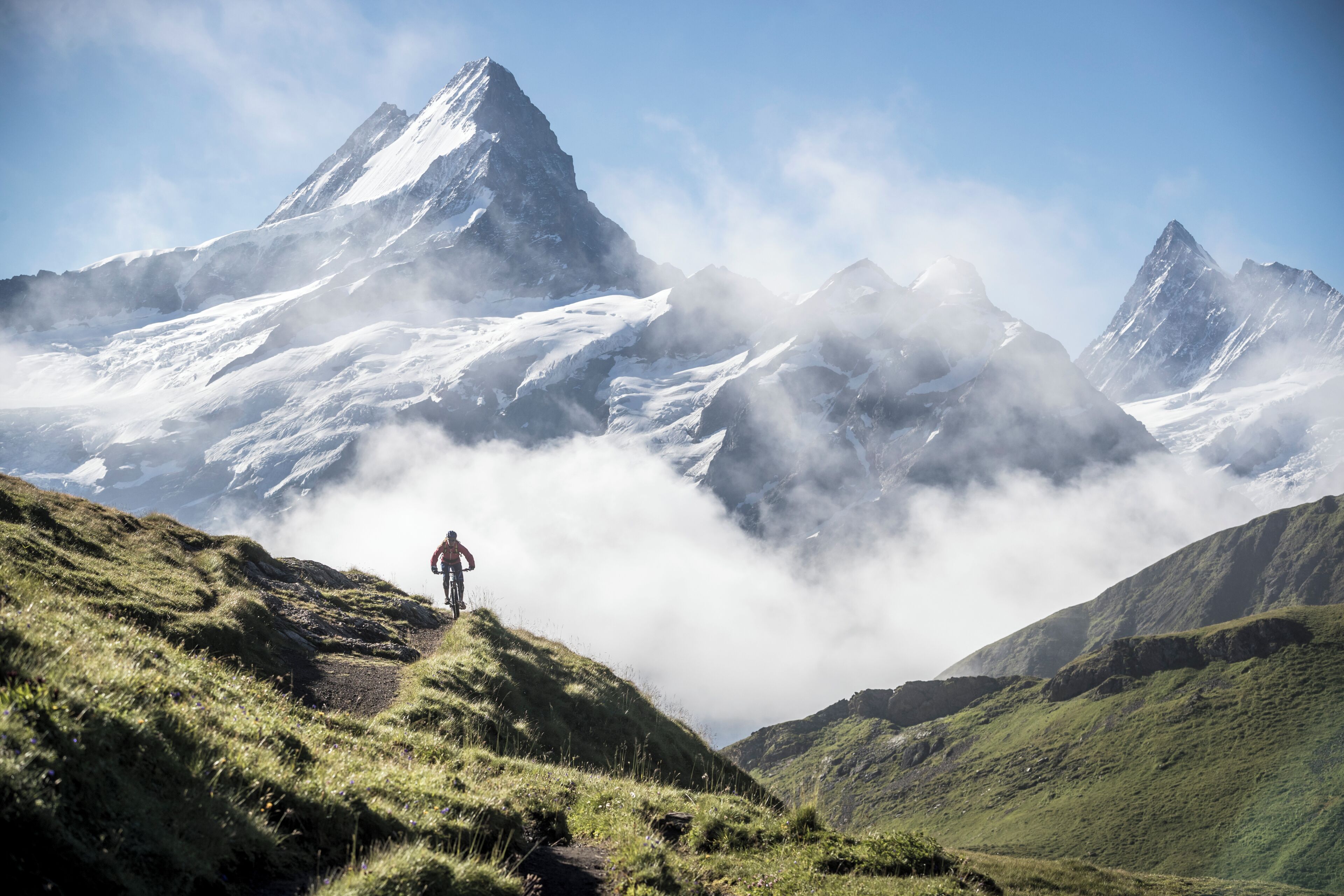A mountain landscape with a person on a trail, snow-covered peaks, and a clear sky.