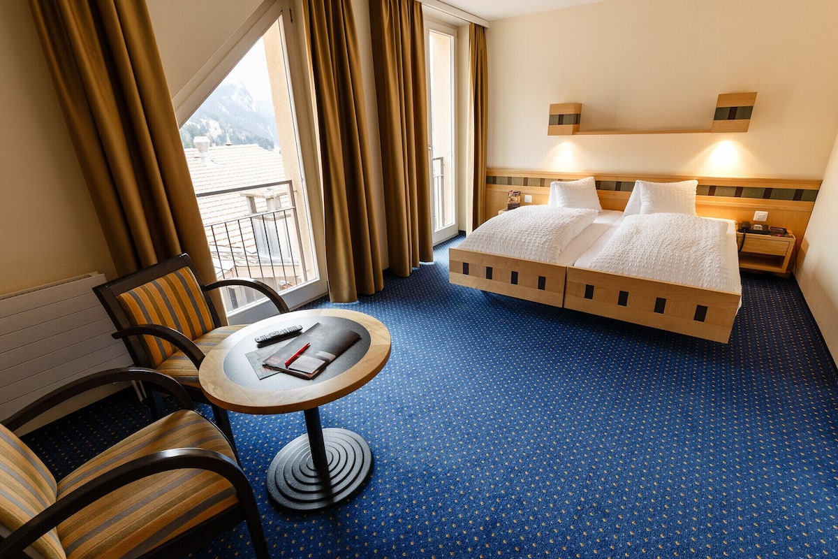 Ein Hotelzimmer mit Bett, einem kleinen Tisch mit Stuhl, einem Fenster mit Vorhängen und Blick auf Berge.