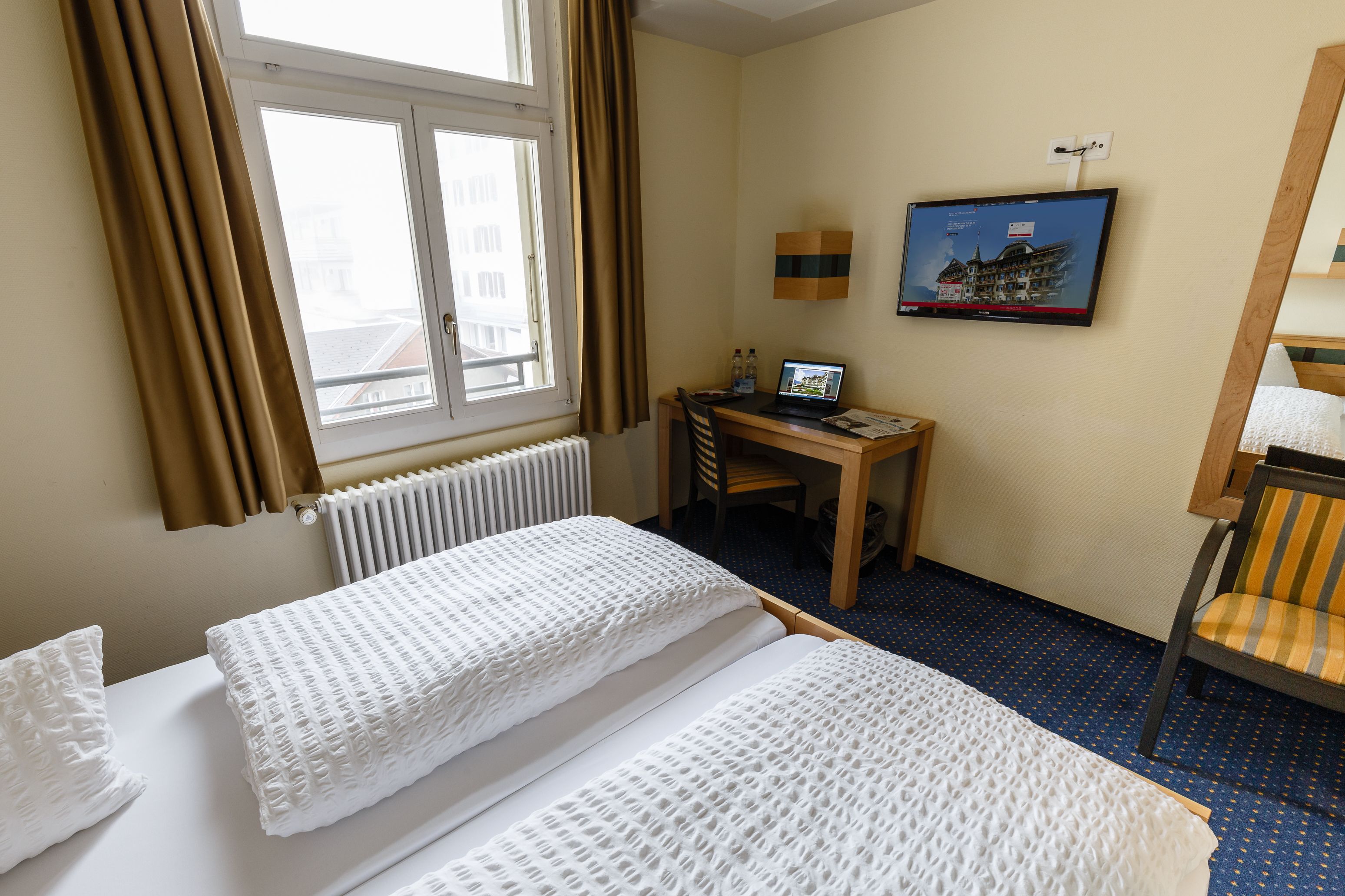 Et hotelværelse med to senge, et skrivebord med en bærbar computer, et fjernsyn på væggen og et vindue med gardiner.
