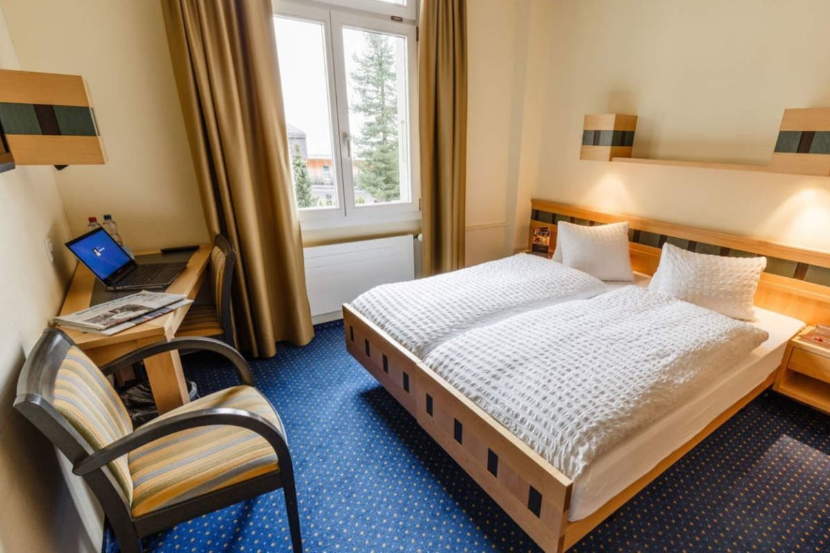 Hotellihuone, jossa on sänky, työpöytä kannettavalla tietokoneella, tuoli ja ikkuna, jossa on verhot.