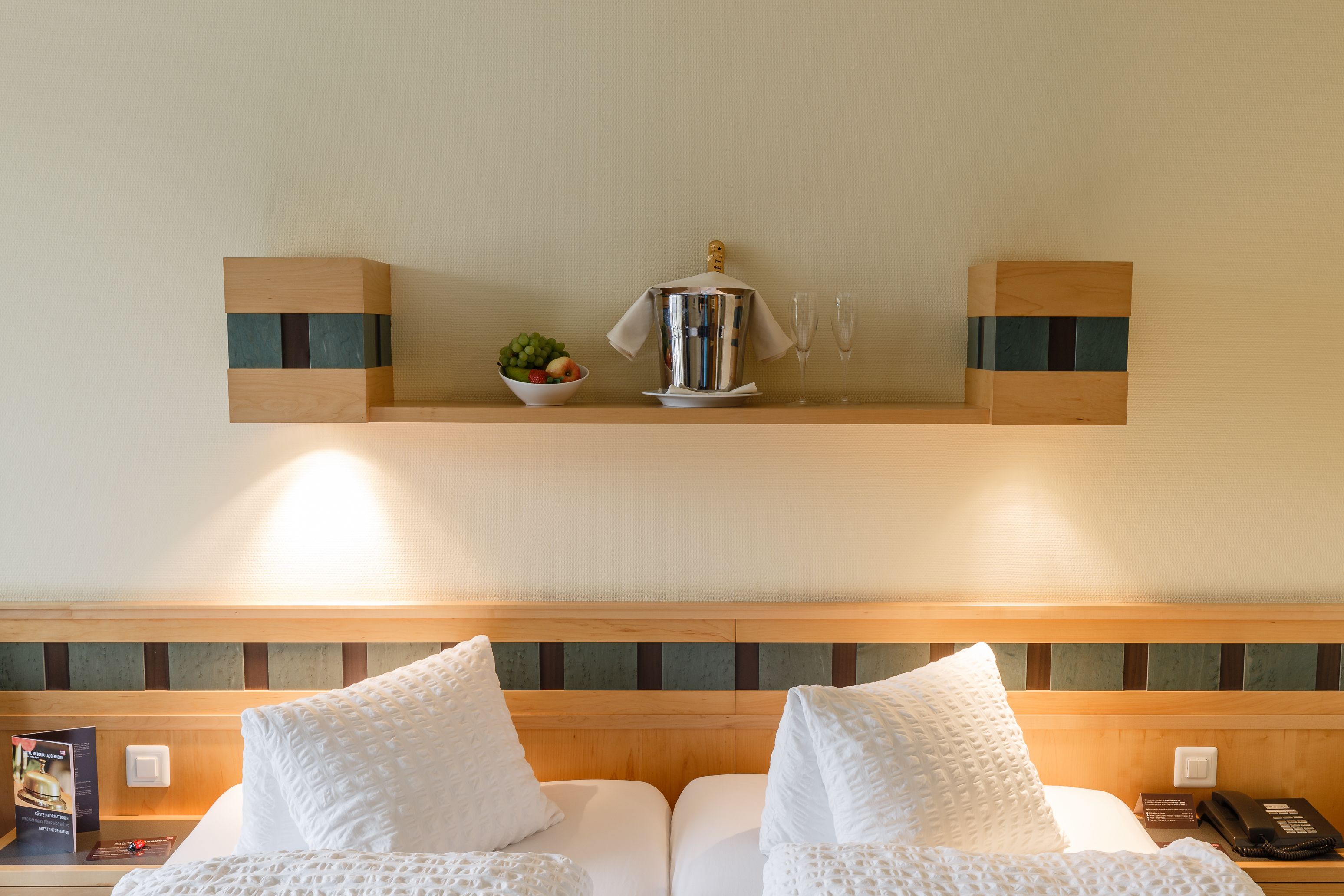Una habitación de hotel con cabecera de madera, dos almohadas blancas, un minibar con una botella y vasos, y un frutero pequeño.