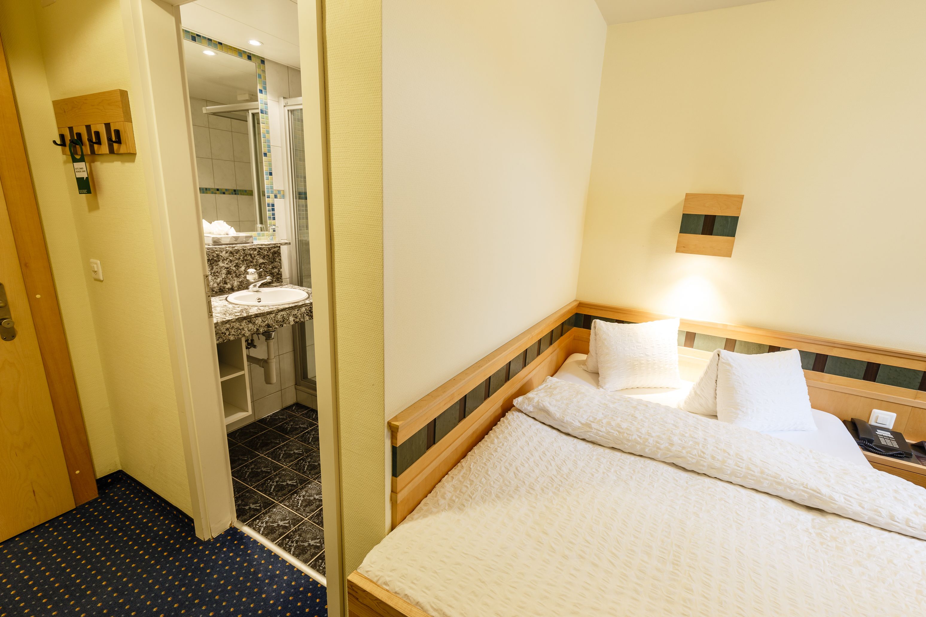 Hotelkamer met een bed, nachtkastje met telefoon, wandlamp, badkamer met wastafel en spiegel, en een deur naar een andere kamer.