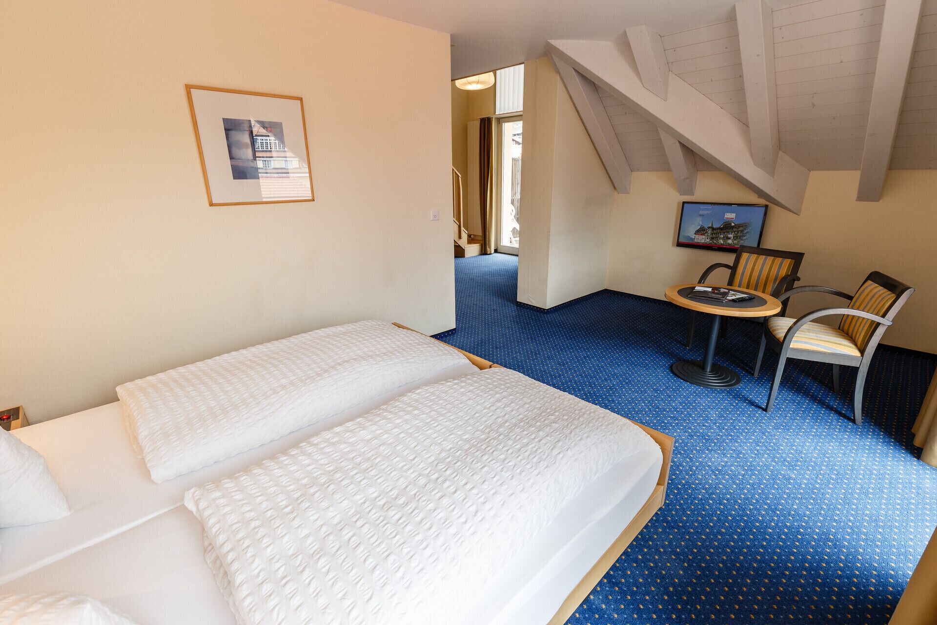 A hotel room with two beds, a small table with chairs, a framed picture on the wall, and a staircase leading to another room.