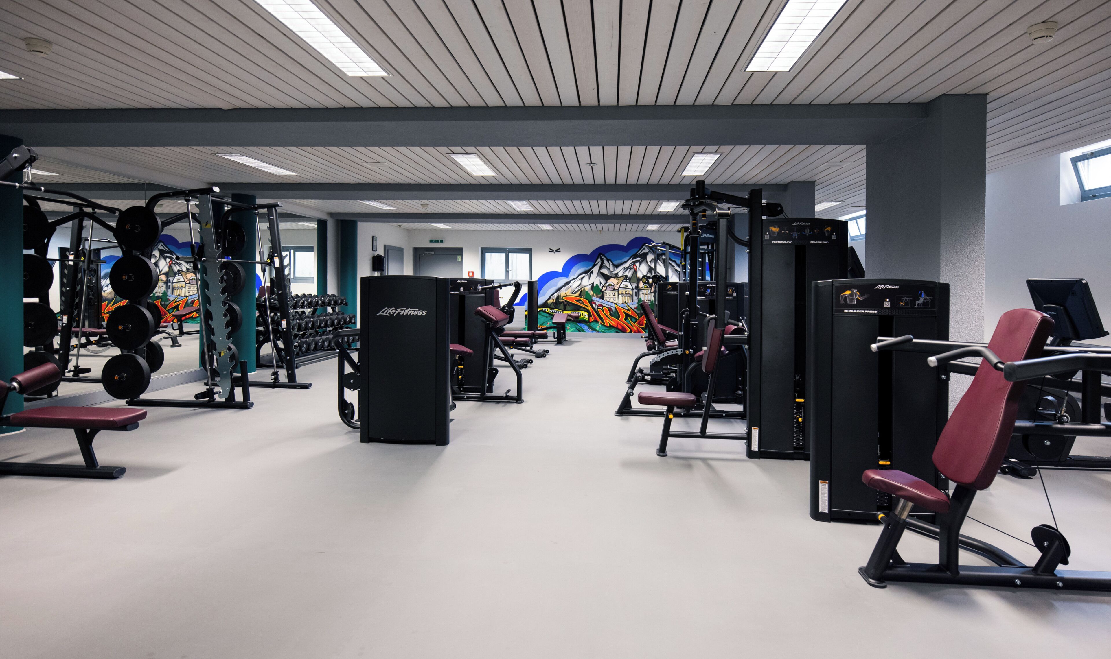 Un gimnasio con diferentes equipos de ejercicio, un mural en la pared y un techo con luminarias lineales.