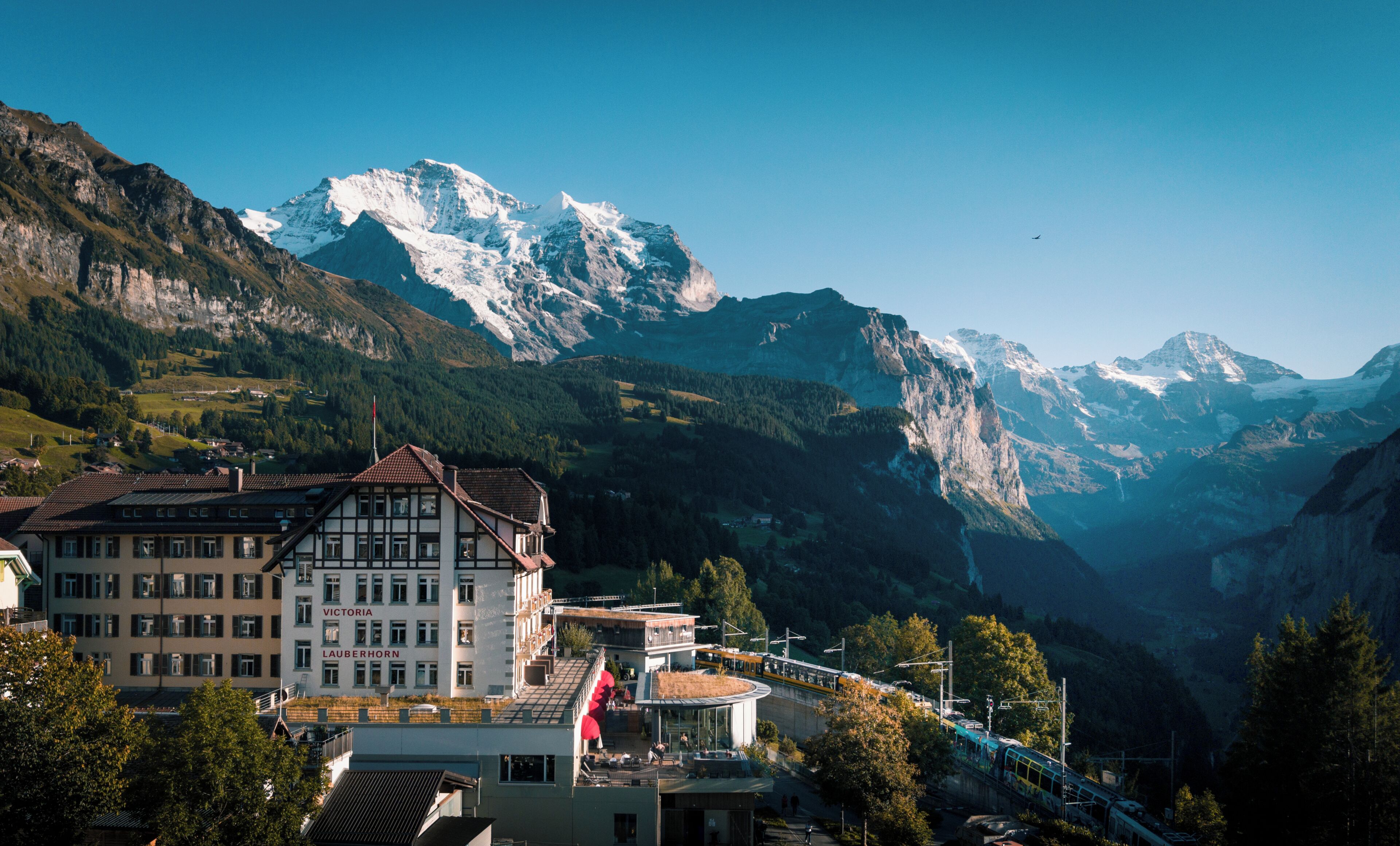 A mountain town with a prominent building featuring a balcony and a flag, surrounded by snow-capped mountains and a train track.