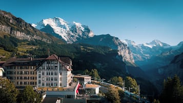 A mountain town with a prominent building featuring a balcony and a flag, surrounded by snow-capped mountains and a train track.