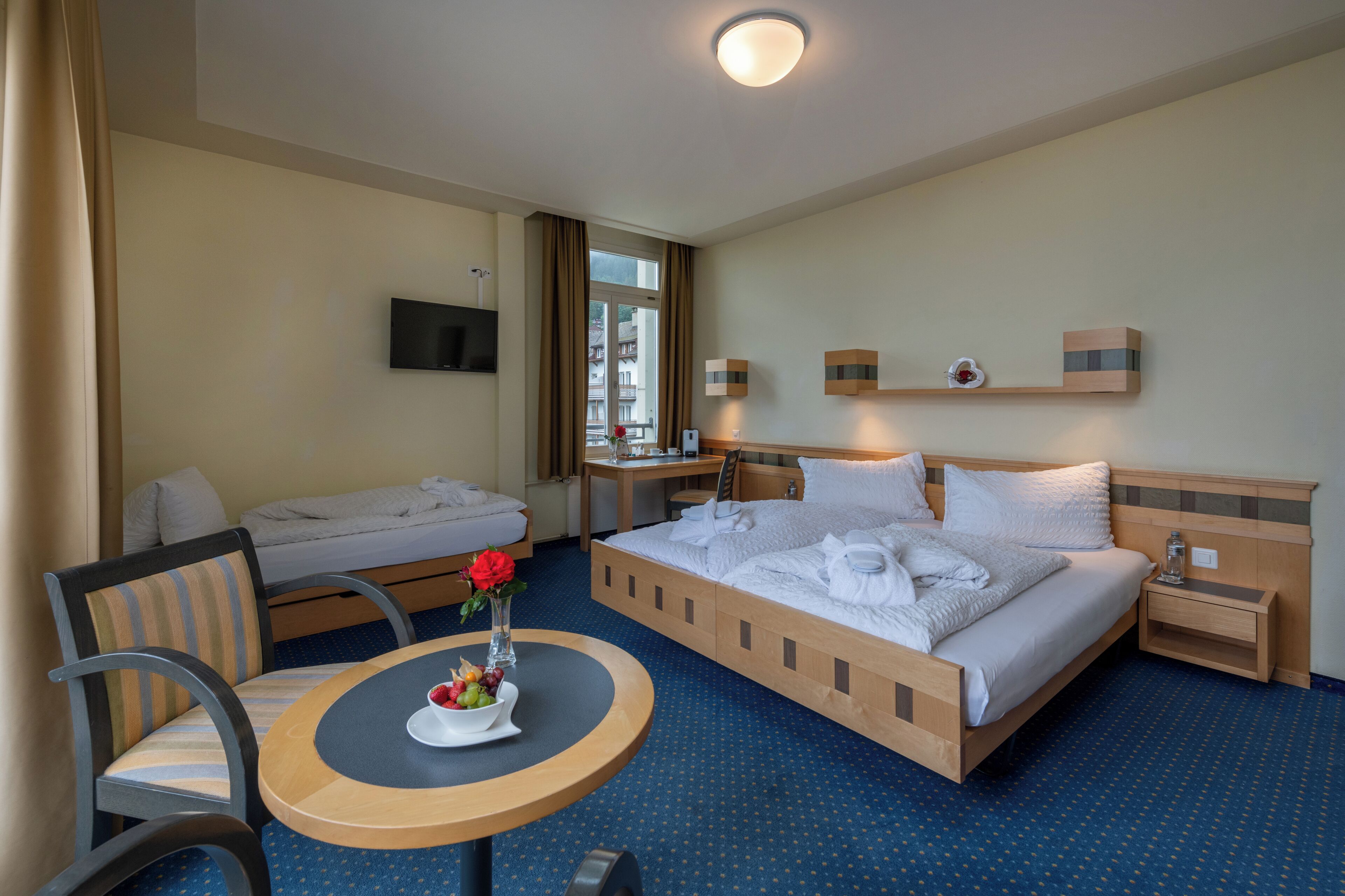 Una camera d'albergo con un letto, una scrivania, una sedia, un tavolino con un vaso di fiori e una televisione appesa al muro.