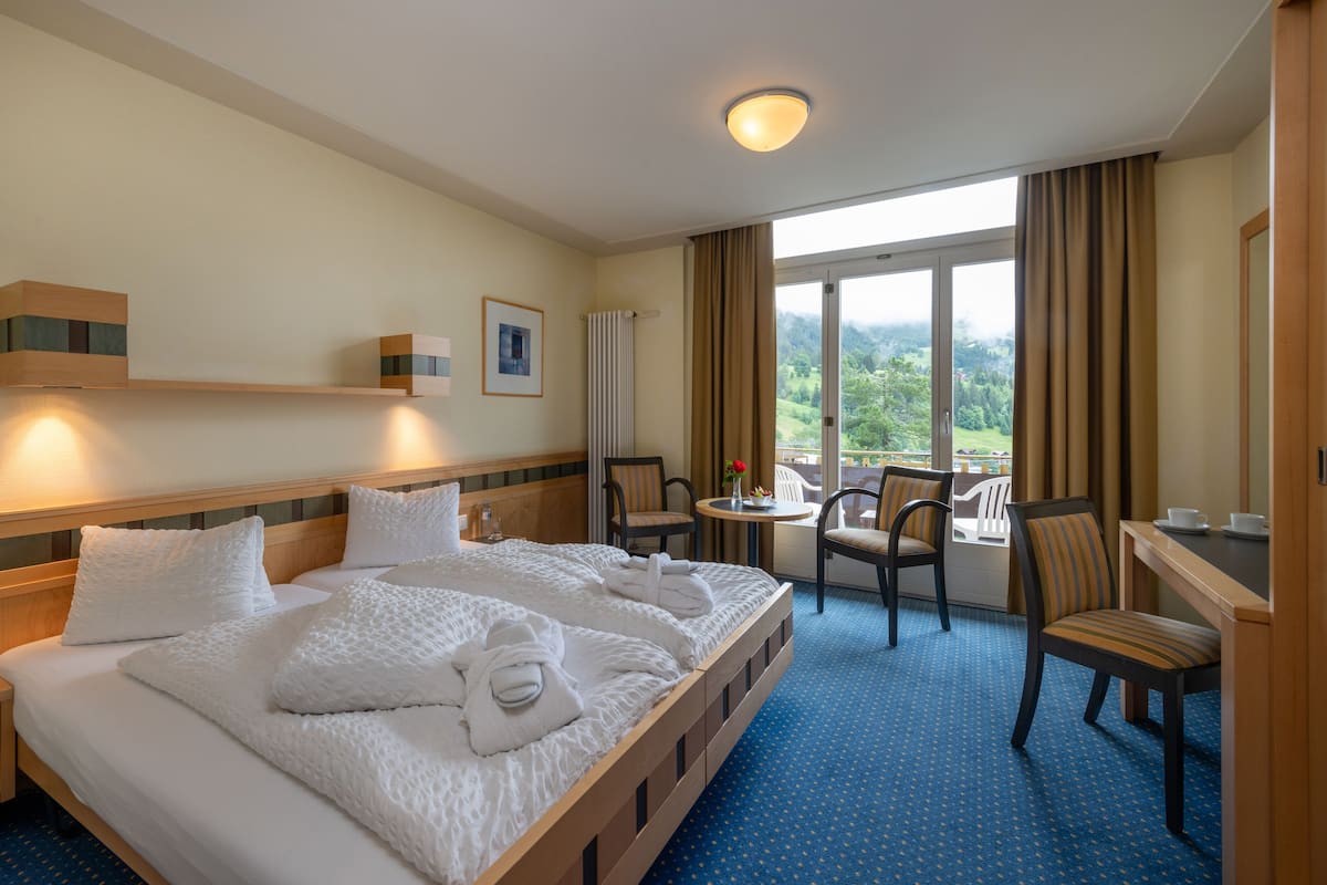 Ein Hotelzimmer mit einem großen Bett, einem Schreibtisch und einem Stuhl. Ein Fenster bietet Blick auf Grünfläche.
