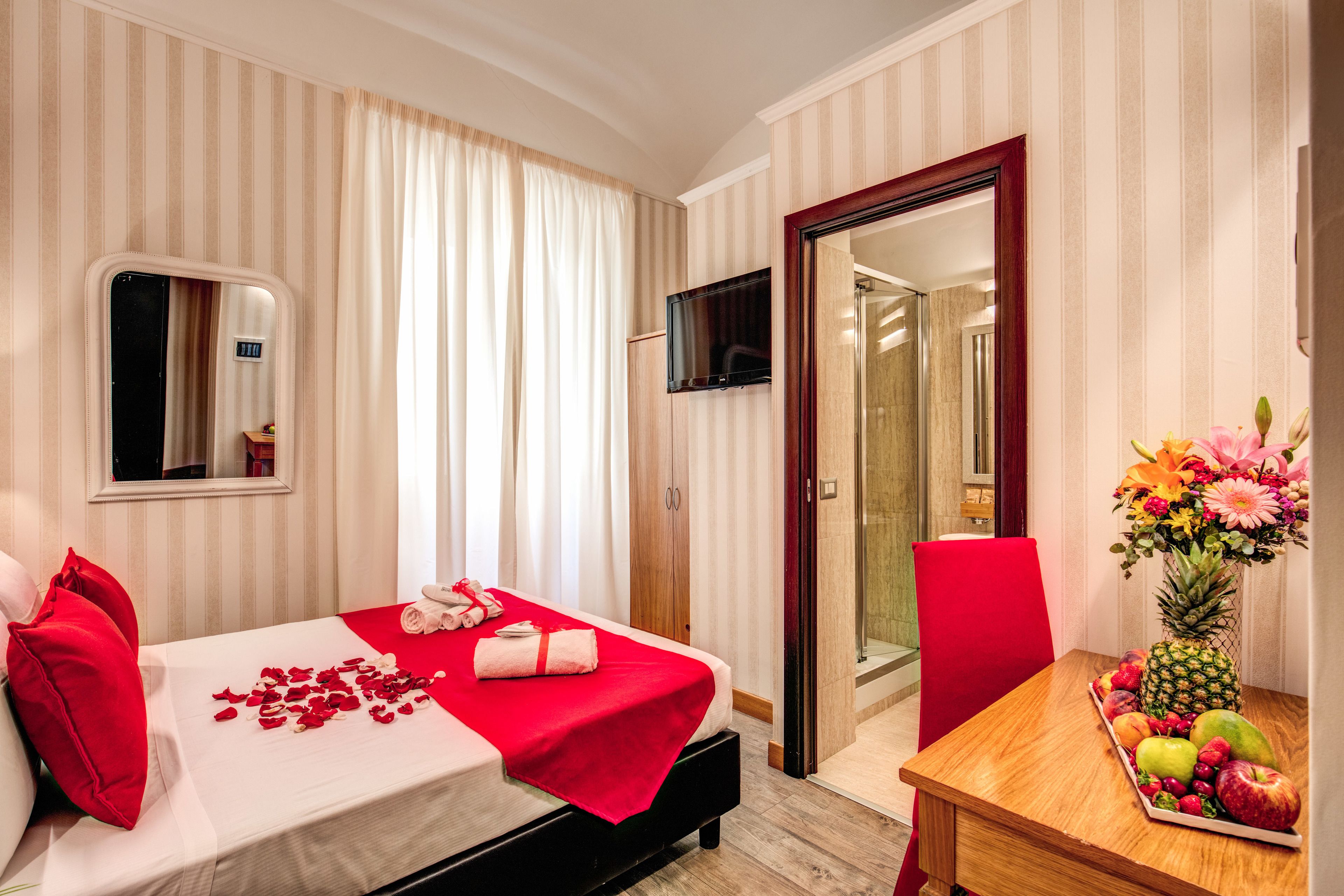 A hotel room with a bed, a table with fruit and flowers, a TV, and a mirror.