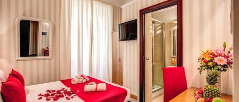 A hotel room with a bed, a table with fruit and flowers, a TV, and a mirror.