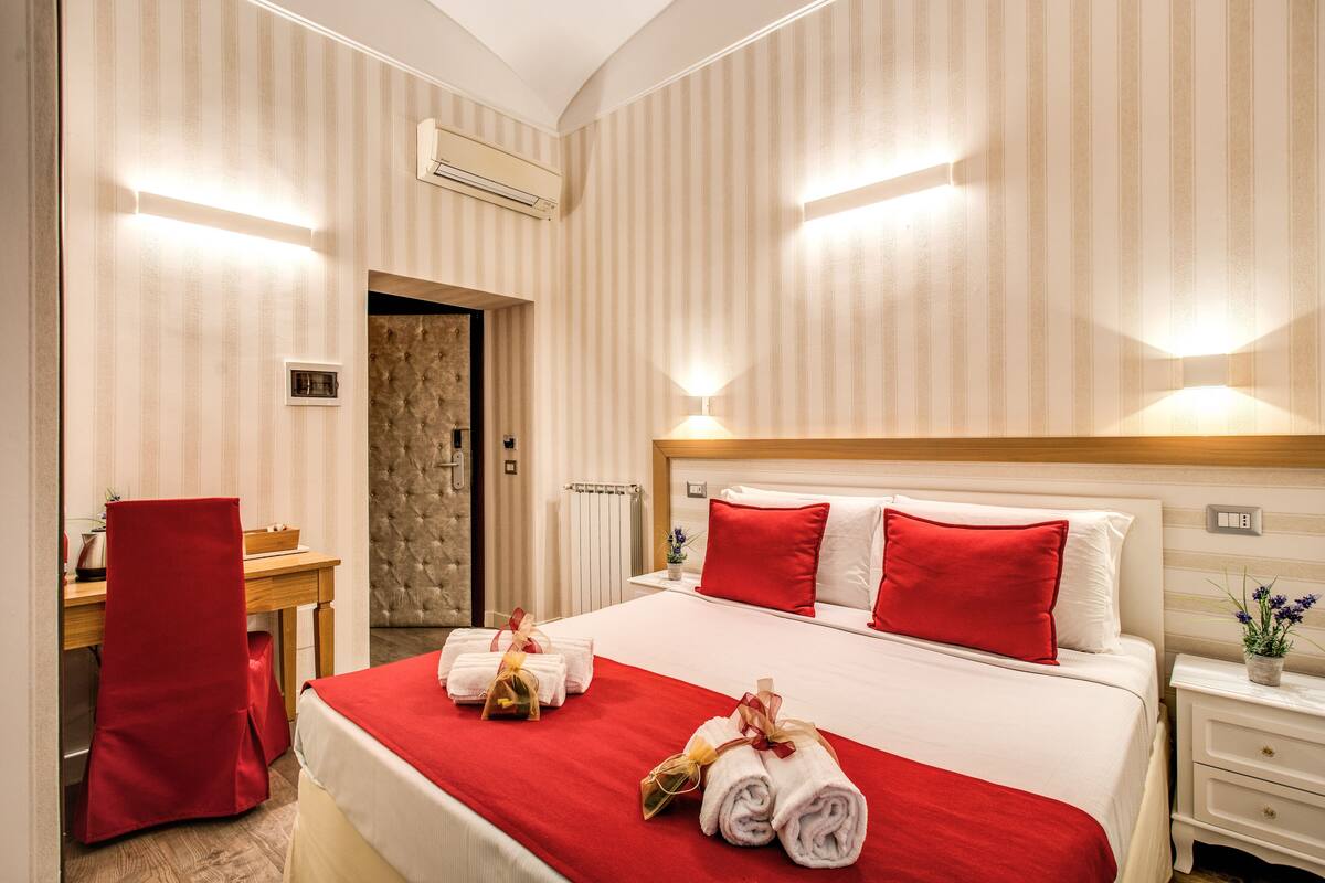 Une chambre d’hôtel avec un grand lit, une literie rouge et blanche, un bureau avec une chaise et une table de chevet ornée d’un vase de fleurs.