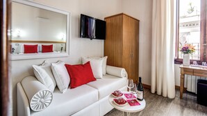 A modern hotel room with a white sofa, red pillows, a wooden wardrobe, a flat-screen TV, a dining table with a vase of flowers, and a window with curtains. - Auditorium Mecenate (Rome)