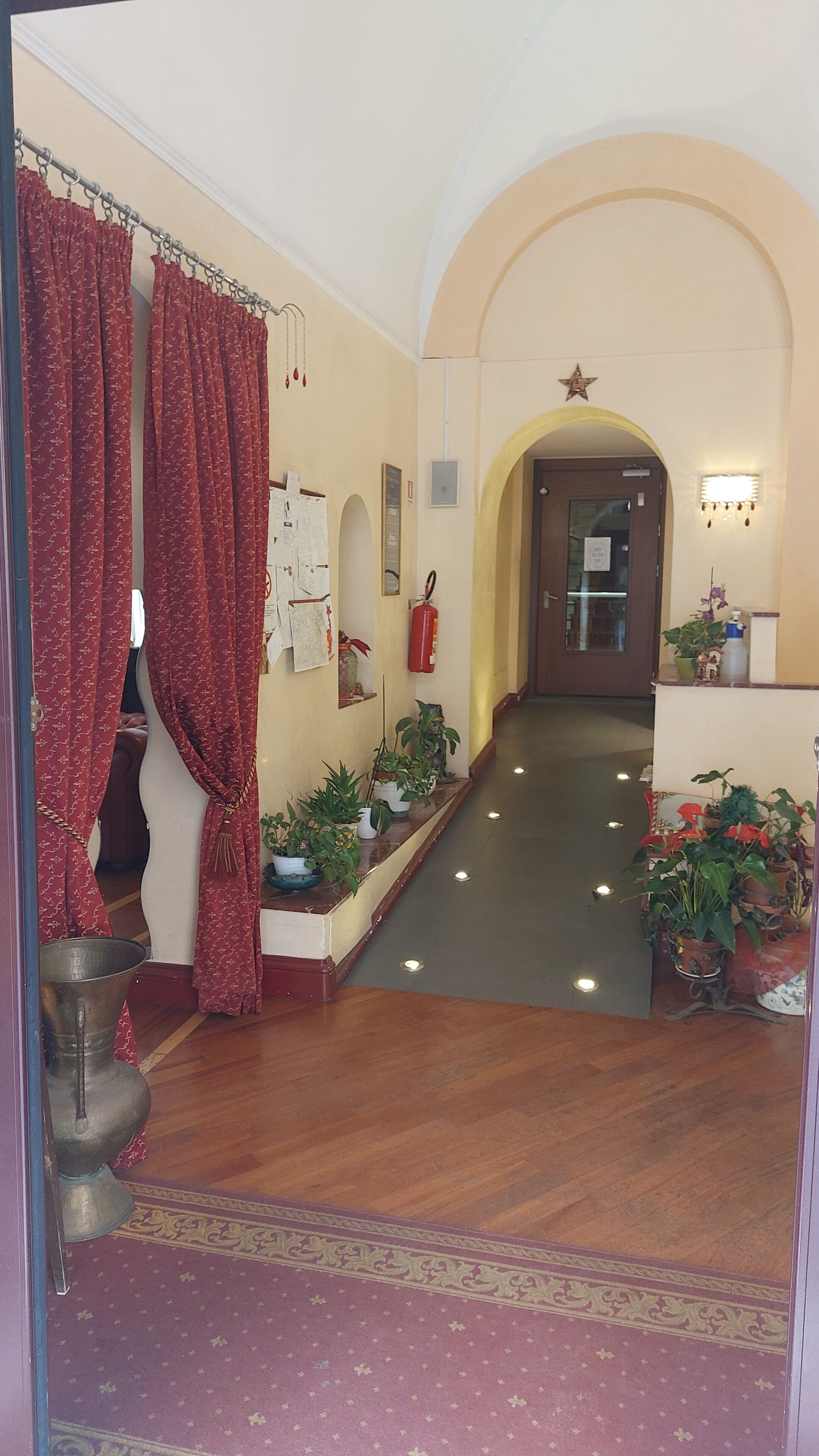 A hallway with a wooden floor, red curtains, potted plants, a fire extinguisher, and a star decoration on the wall.