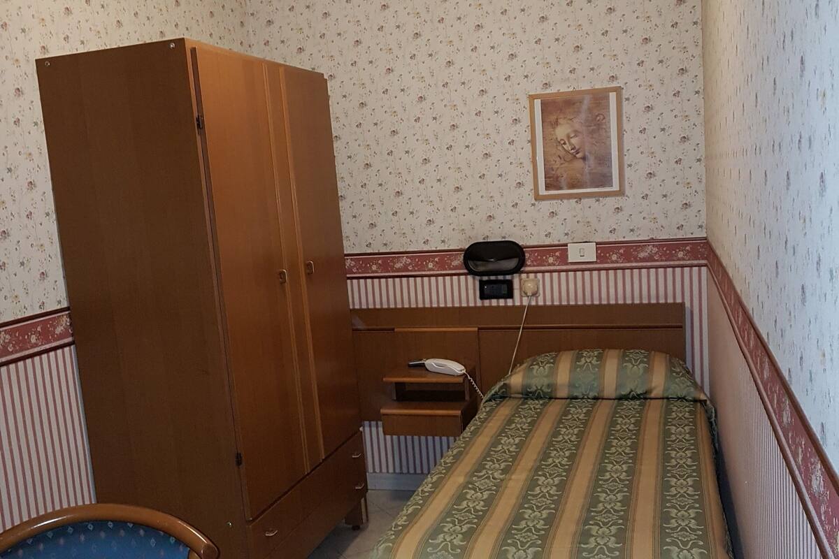 A bedroom with a bed, a wooden wardrobe, a desk with a telephone, and a chair.
