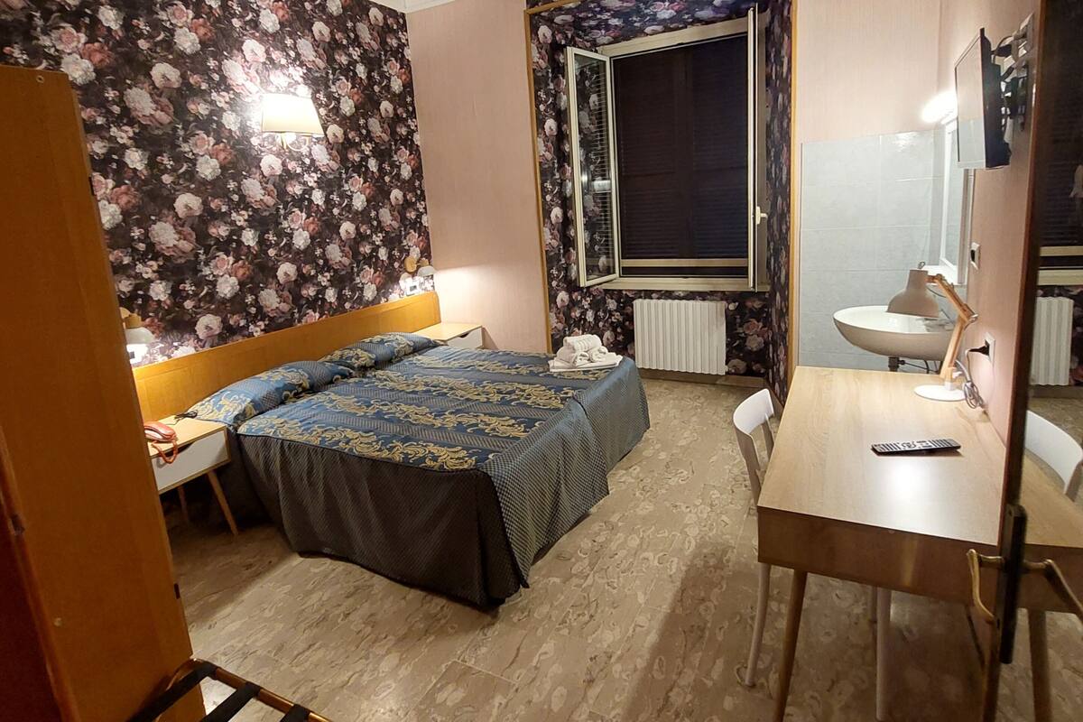 A hotel room with a bed, a desk, a chair, a window, and a radiator.