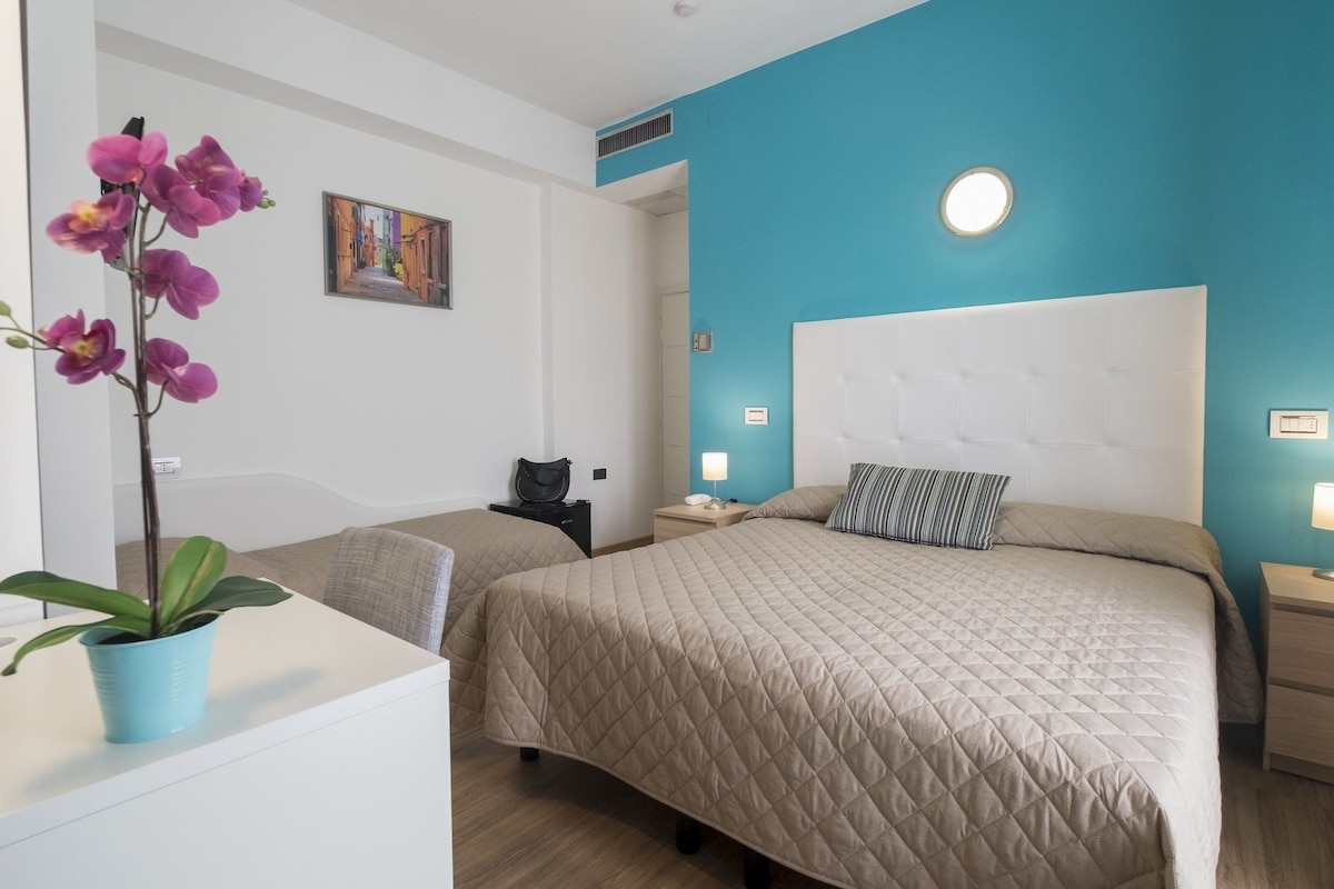 A hotel room with a large bed, a smaller bed, a nightstand with a lamp, a wall-mounted air vent, and a potted orchid.