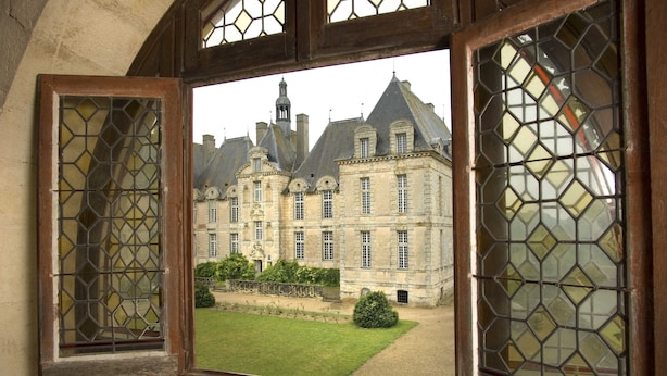 Une vue à travers une fenêtre ornée d’un motif géométrique, donnant sur un grand bâtiment historique doté de nombreuses cheminées et bordé d’une pelouse bien entretenue.