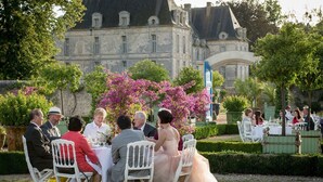 Un espace de restauration en plein air, avec des chaises et des tables blanches, entouré d’une végétation luxuriante et avec un bâtiment historique en arrière-plan.