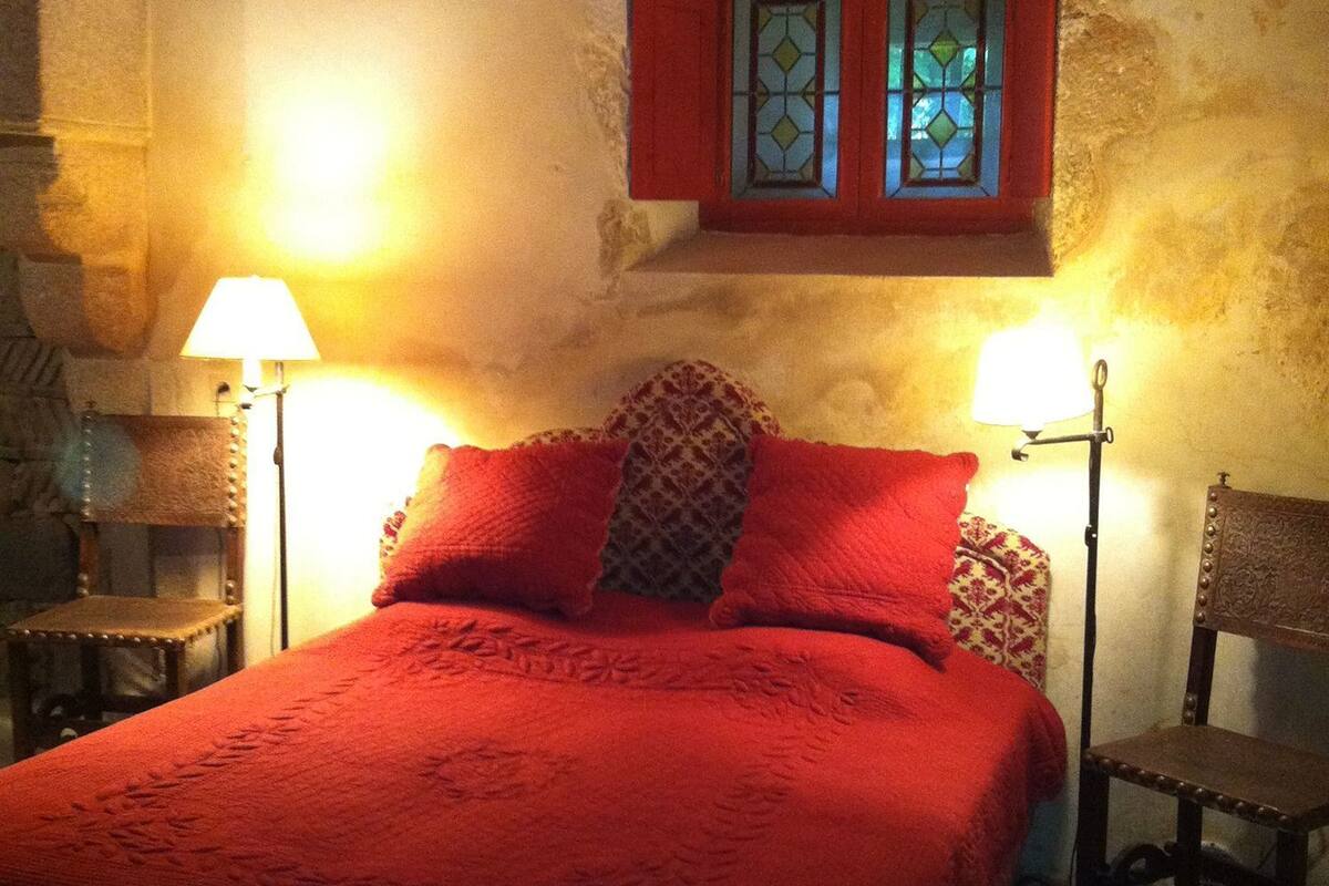 Een bed met een rode sprei, twee nachtlampjes en een raam met glas-in-lood.