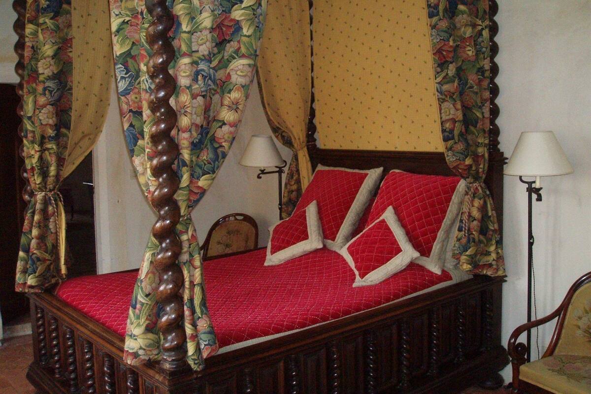 Een hemelbed met bloemen gordijnen, een rode geborduurde sprei en twee bedlampen.