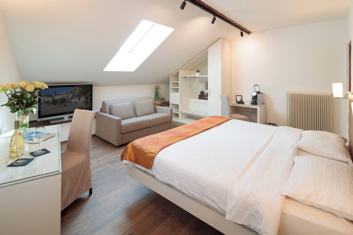 A modern hotel room with a bed, a sofa, a desk with a computer, and a TV.