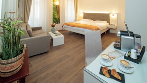A modern hotel room with a bed, a sofa, a coffee table, a dining table with croissants and coffee cups, and a coffee machine. - Hotel America (Locarno)