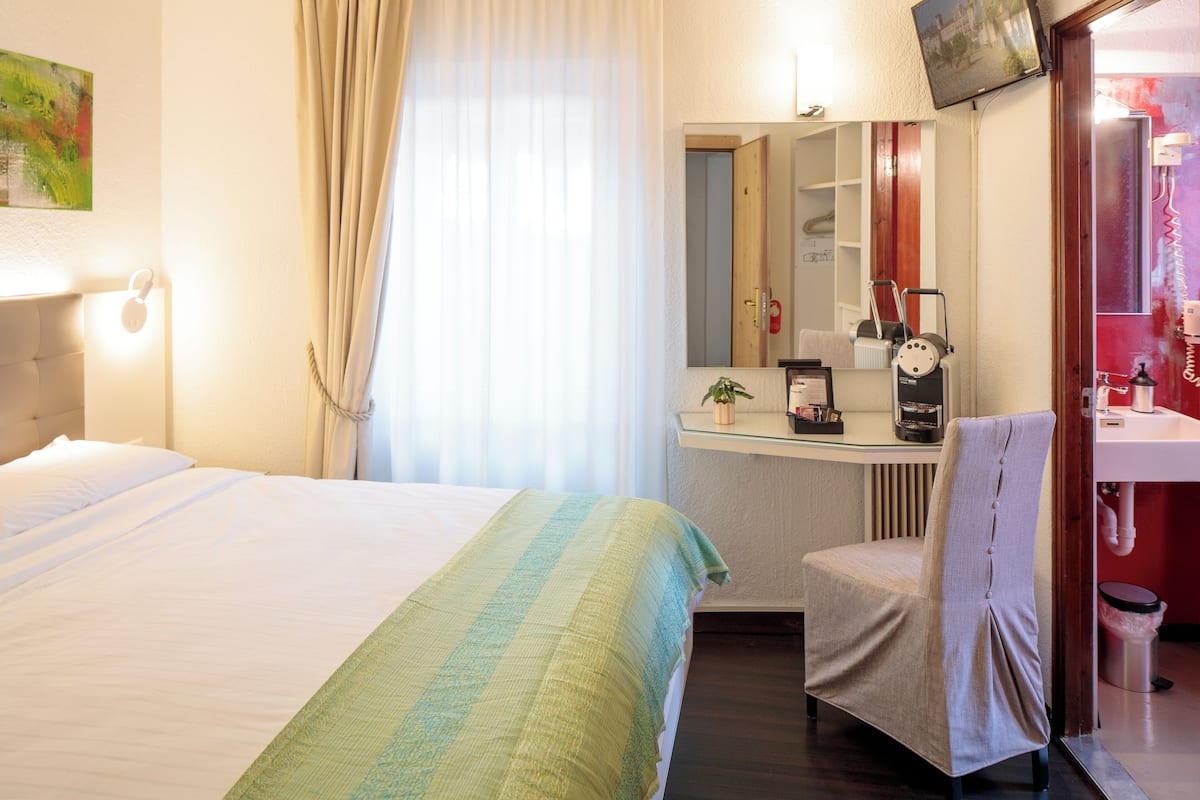 A hotel room with a bed, a desk with a chair, a mirror, a sink, and a trash can.