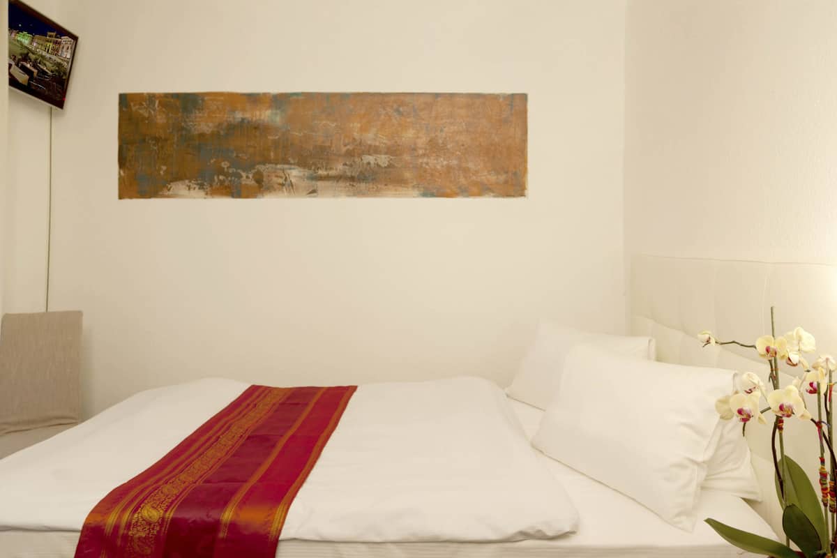 A hotel room with a large bed, a red and gold throw blanket, white pillows, and a single orchid plant.