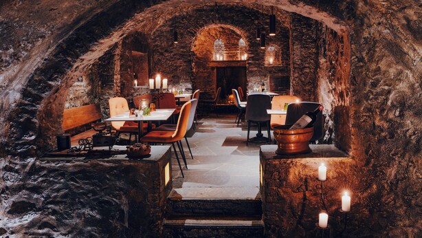 An underground dining area with stone walls, tables, chairs, and candles. - Hotel America (Locarno)