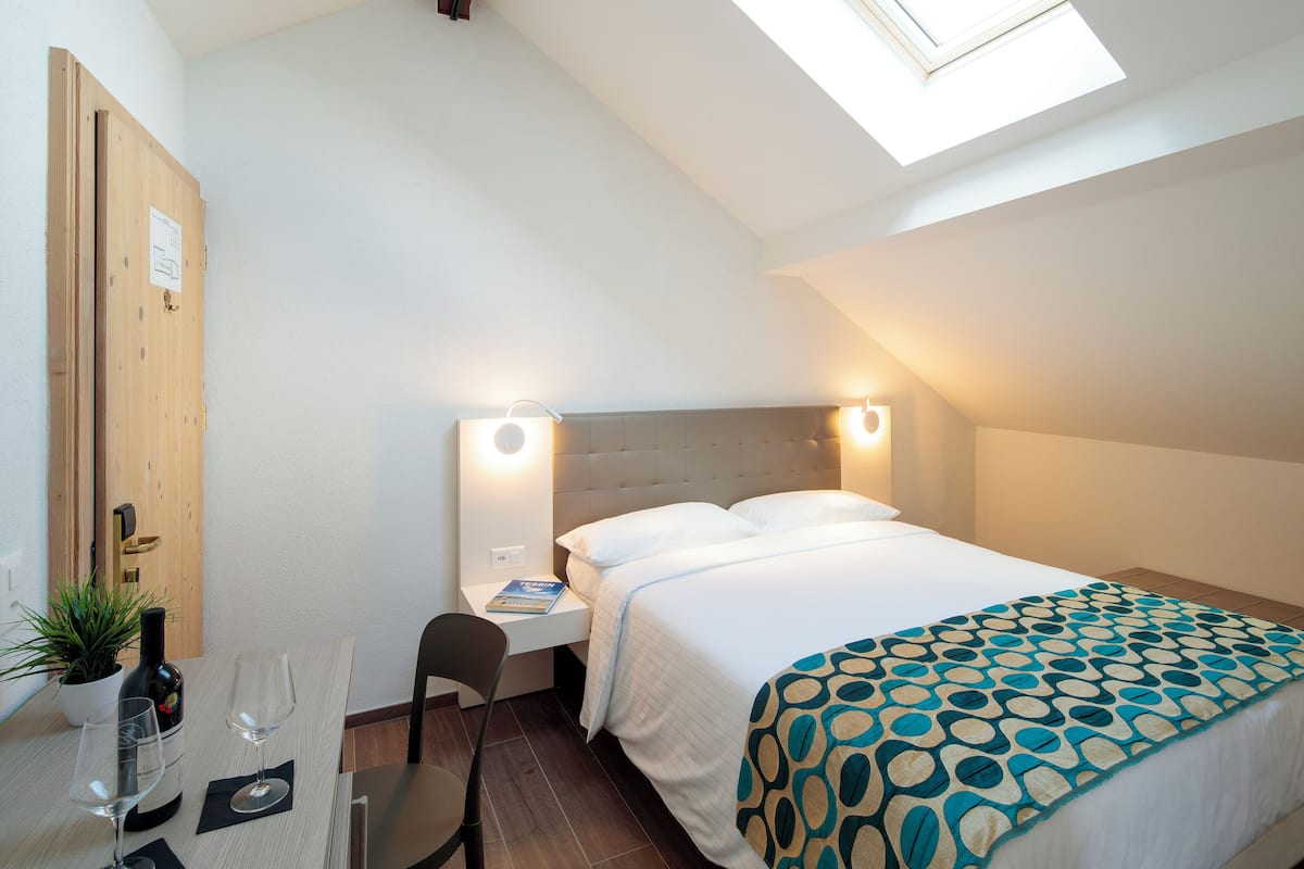 A hotel room with a bed, bedside table, chair, and a small table with a bottle and glasses.