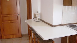 A small kitchen with a white countertop, wooden stools, a coffee maker, and a stove. - Popi (Chania)