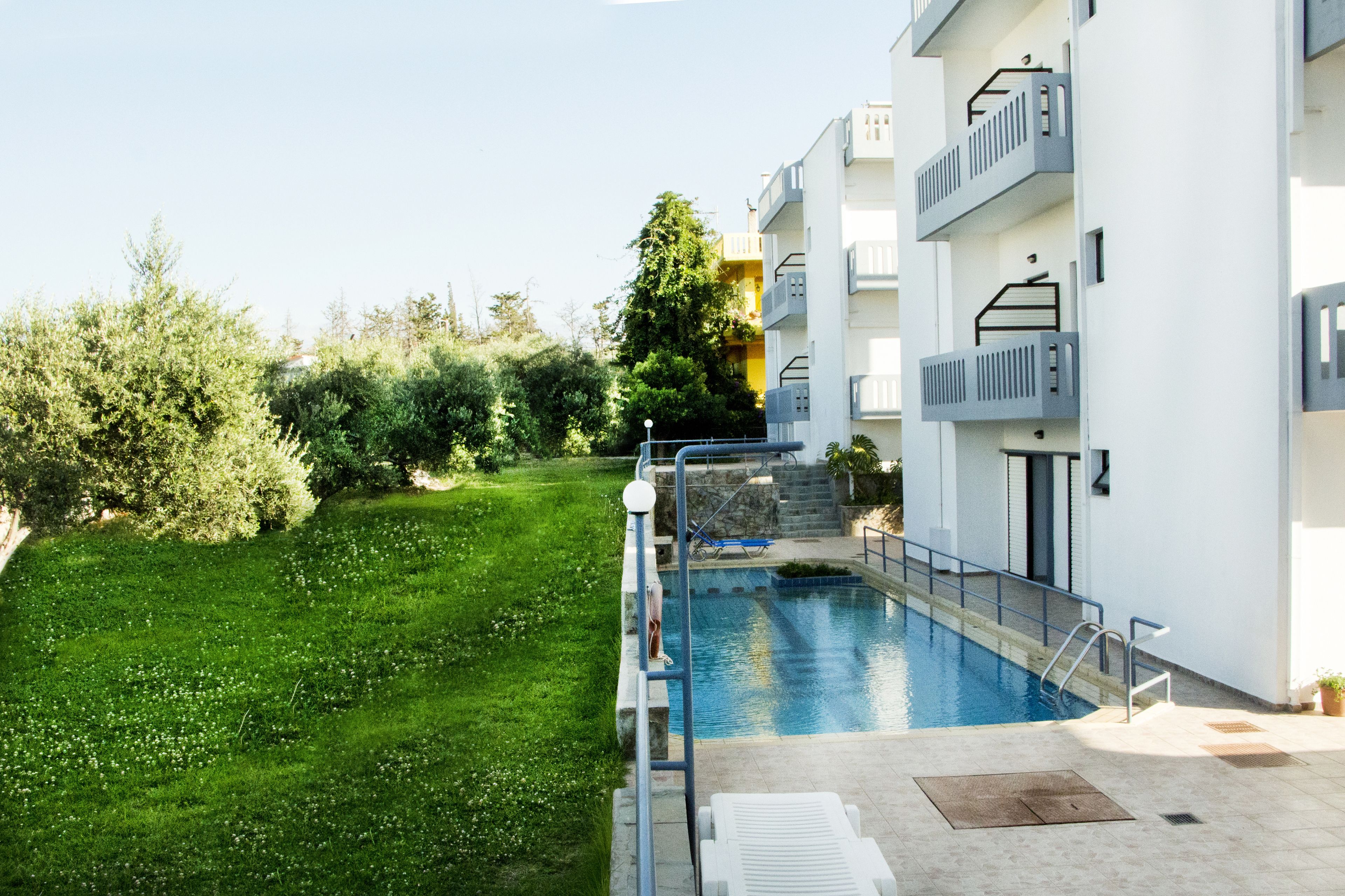 Piscina contigua a un edificio moderno con balcones, rodeada de vegetación.