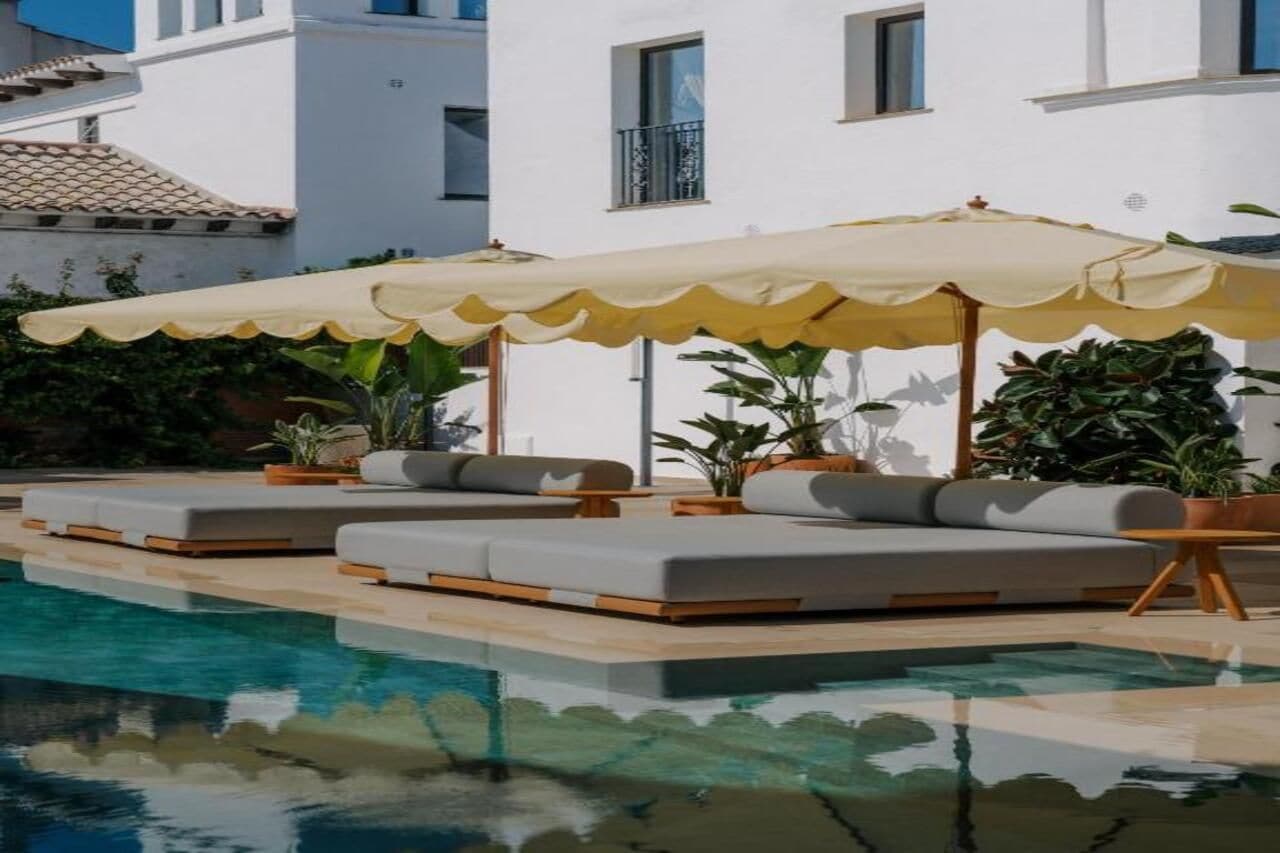 Zona de piscina amb gandules i ombrel·les, envoltada d'un edifici blanc amb balcons.
