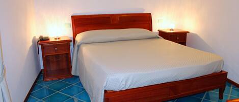 A single-bed room with a wooden headboard, a bedside table with a lamp, and a nightstand with a phone. The room has blue-tiled flooring.