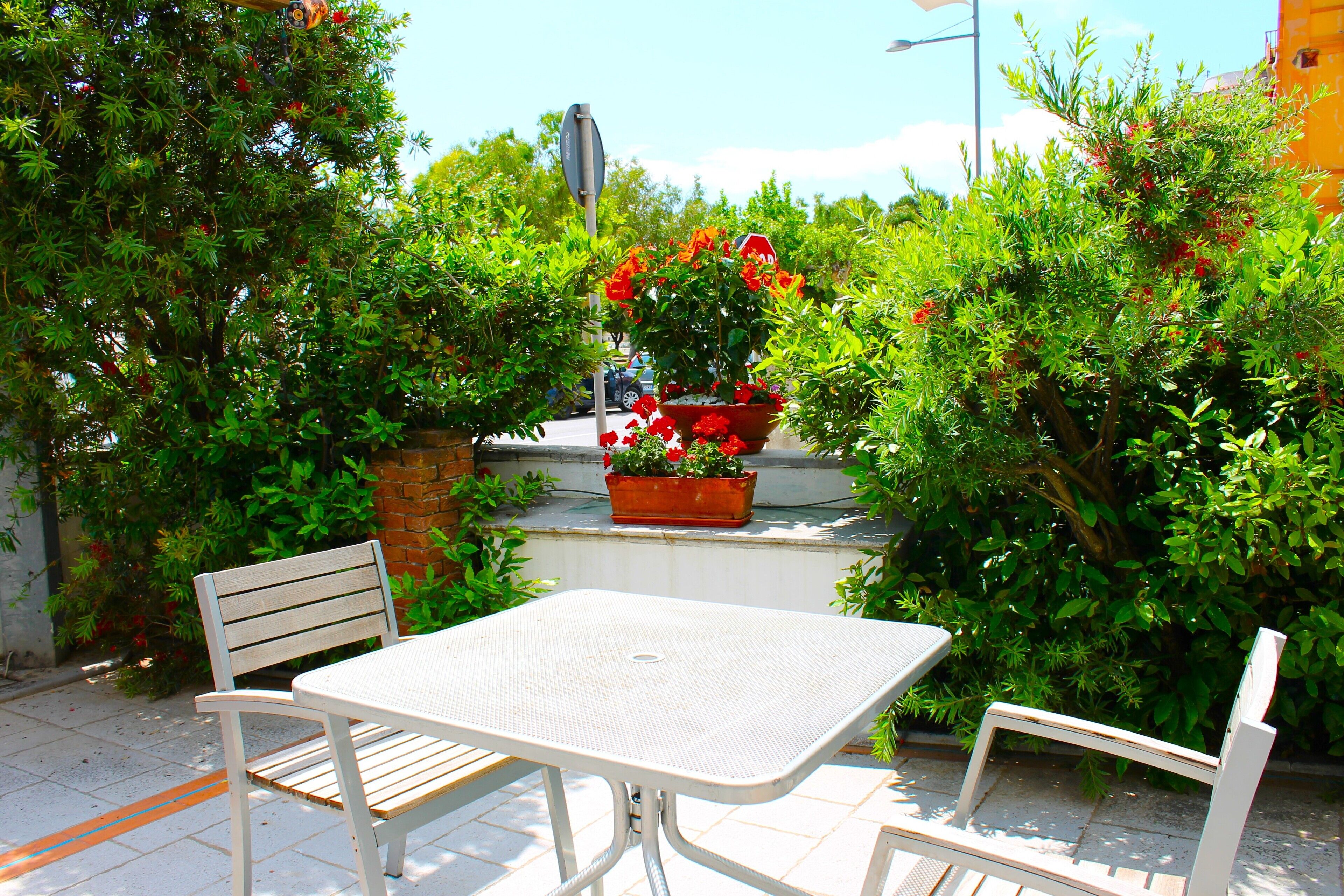 Un patio con un tavolo in metallo bianco e due sedie, immerso nel verde e con una pianta in vaso con fiori rossi.
