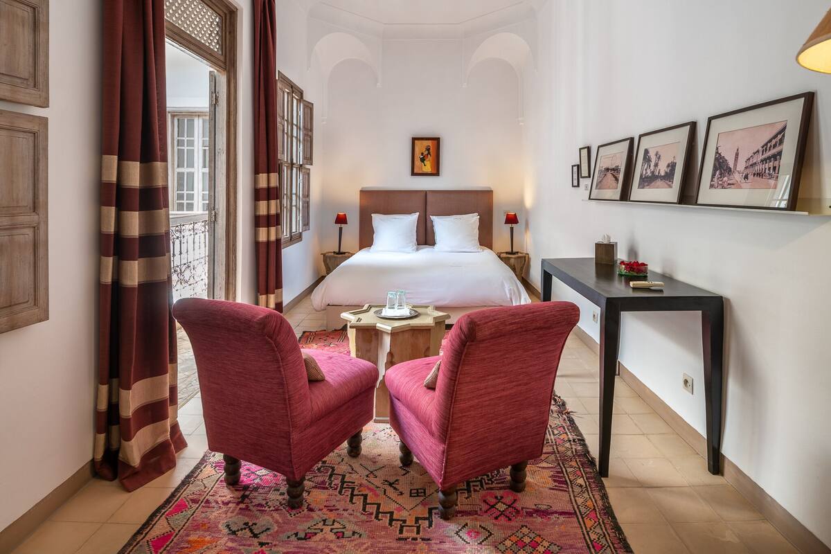A hotel room with a large bed, two red armchairs, a small table, and a desk.