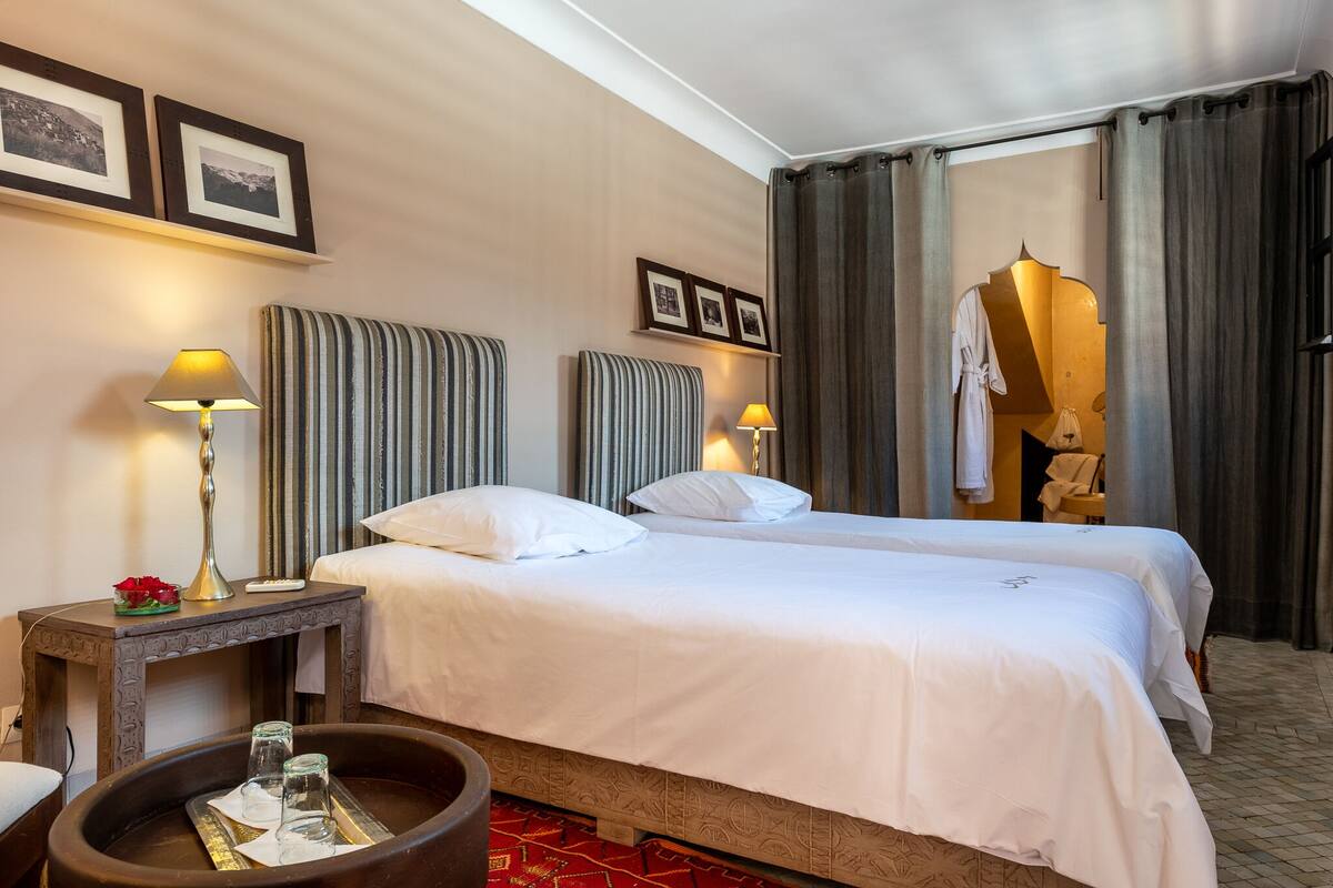 A hotel room with two beds, a small table with a lamp, a decorative bowl, and framed pictures on the wall.