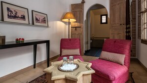 A cozy room with two red armchairs, a wooden side table with glasses, and a lamp. There are framed pictures on the wall and a wooden door in the background.