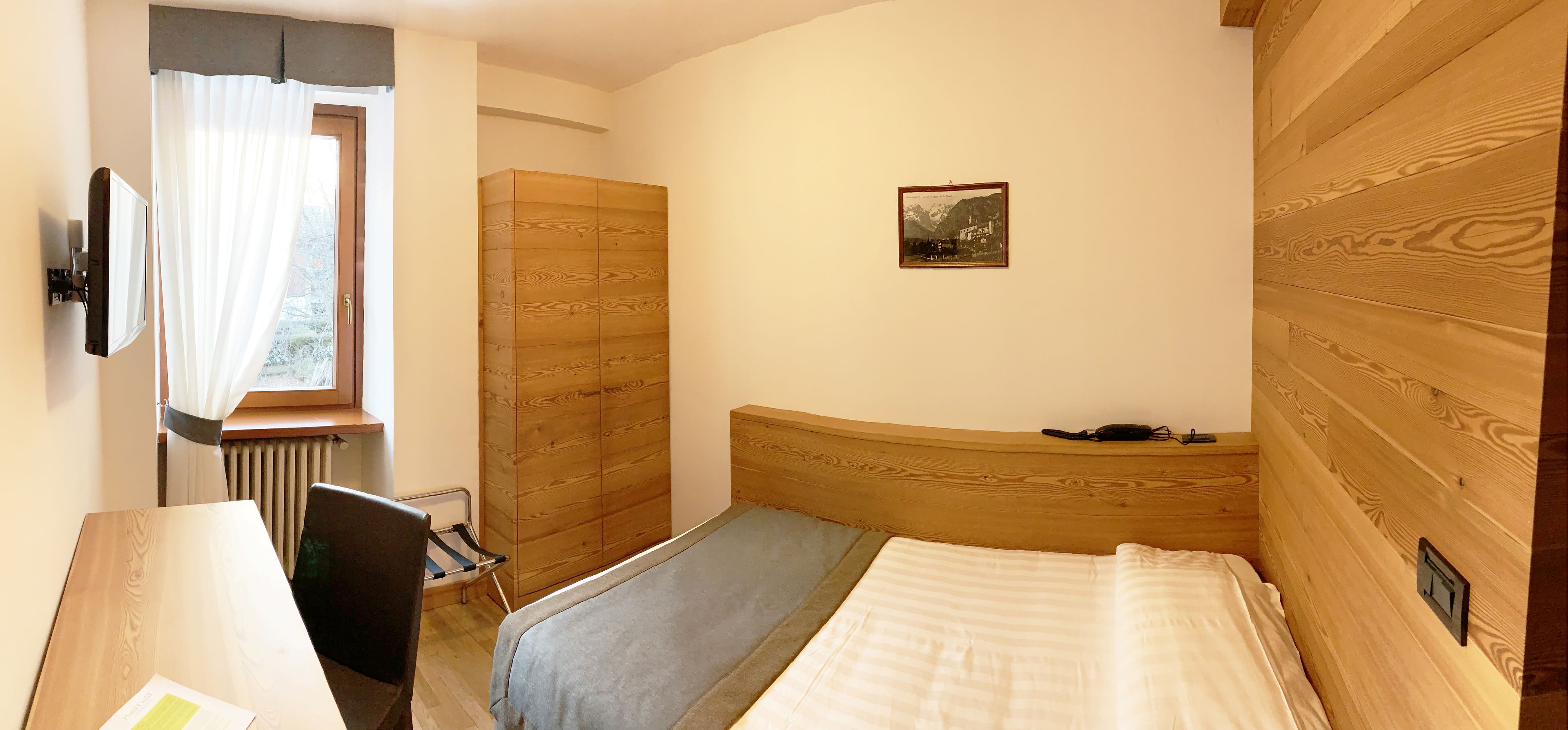 A bedroom with a wooden headboard, a bed, a desk with a chair, a radiator, and a window with curtains.