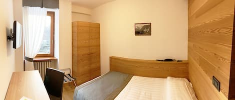 A bedroom with a wooden headboard, a bed, a desk with a chair, a radiator, and a window with curtains.