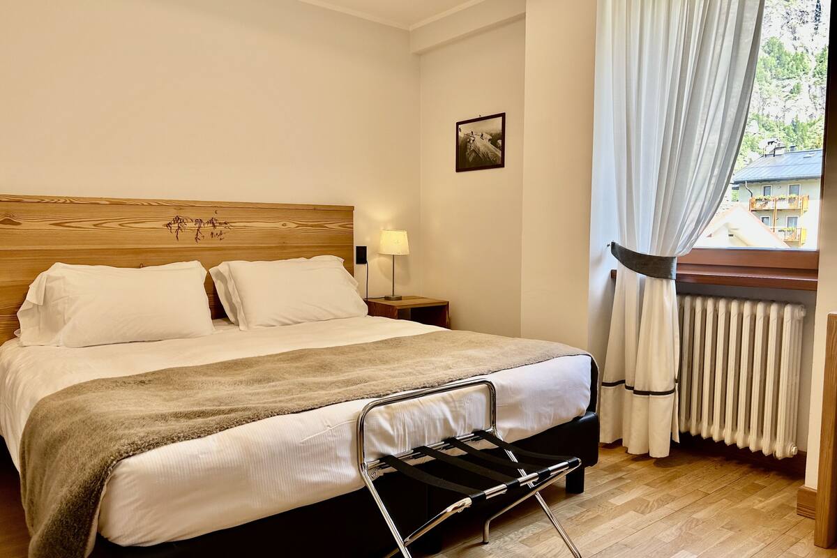 A neatly made bed with a headboard, a bedside lamp, and a view of a building through a window with curtains.