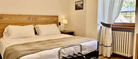 A neatly made bed with a headboard, a bedside lamp, and a view of a building through a window with curtains.