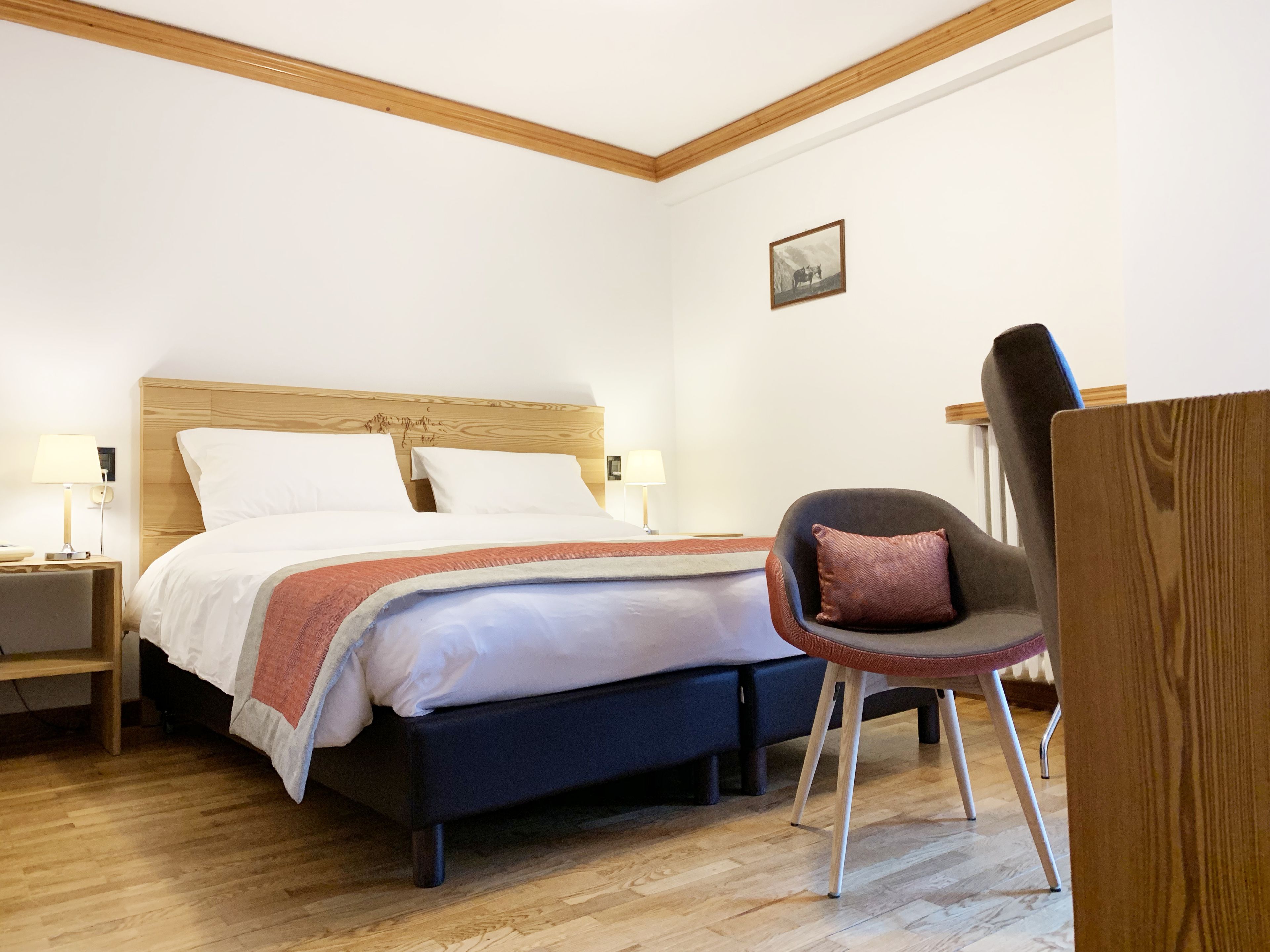 A bedroom with a wooden headboard bed, two bedside lamps, a chair with a cushion, and a small picture on the wall.