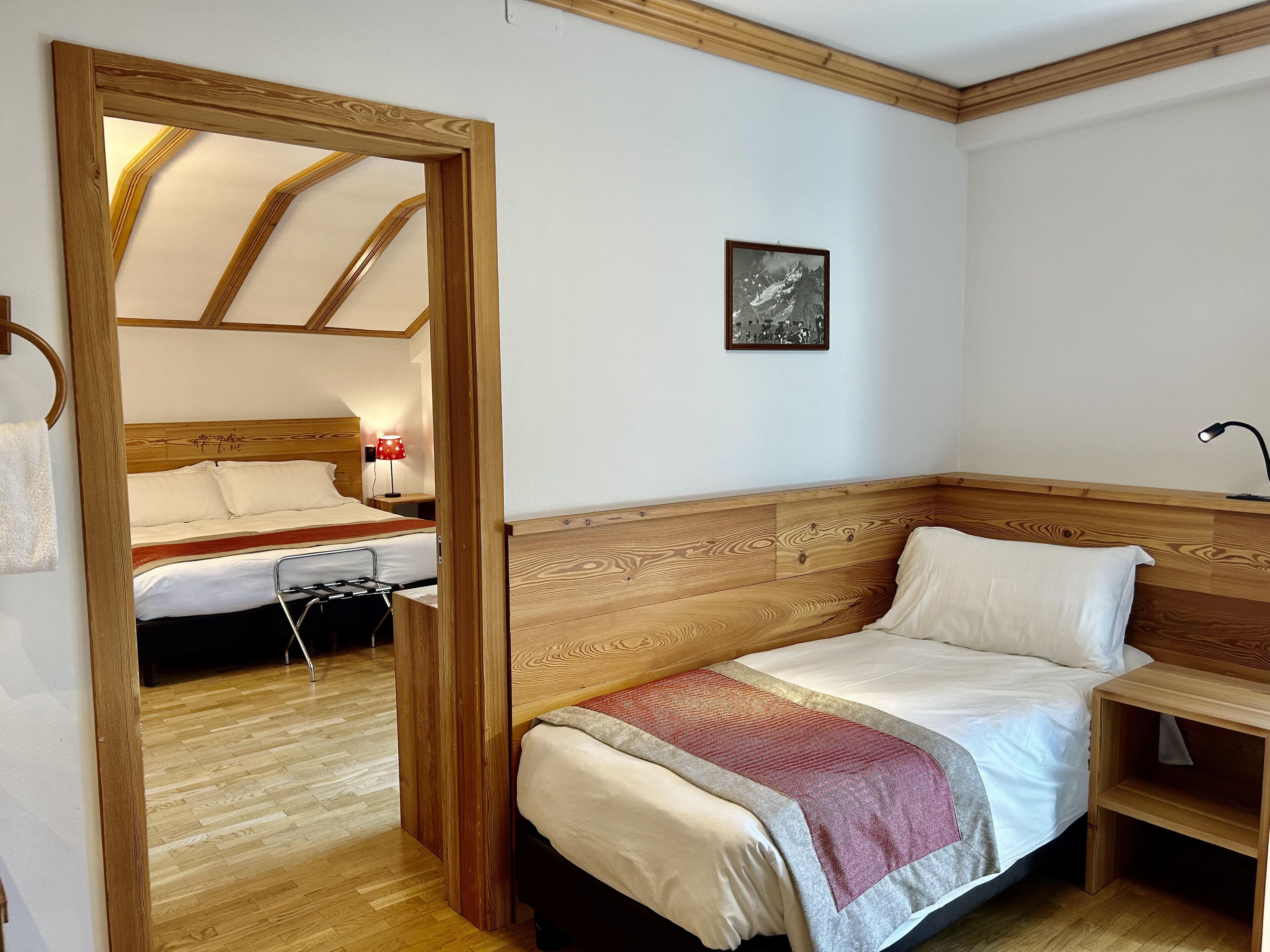 Una habitación con cabecera de madera, cama con manta roja y gris, mesita de noche con lámpara y un cuadro enmarcado en la pared.