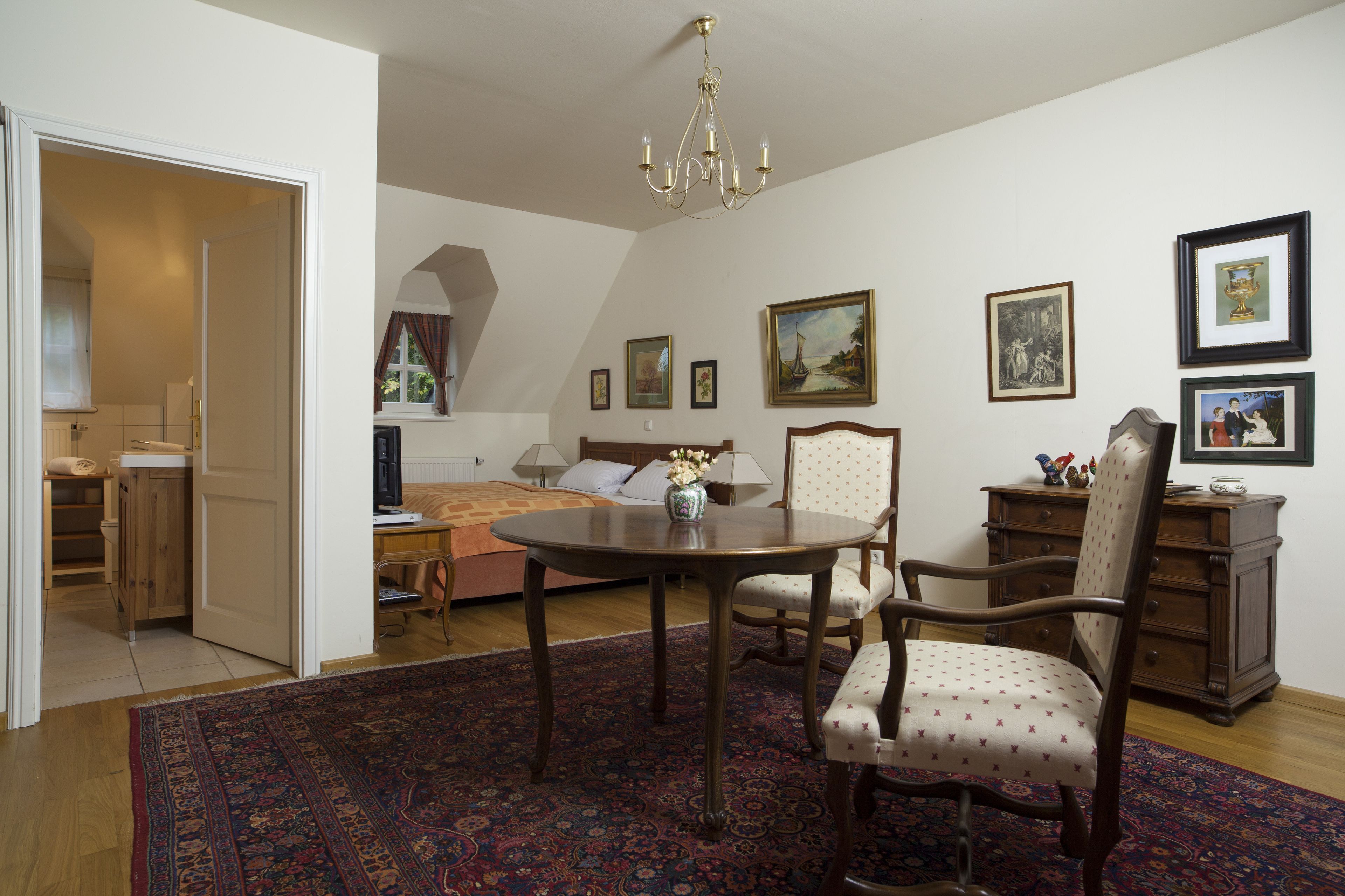 A room with a round wooden table, a chair, a bed, a dresser, and a chandelier.
