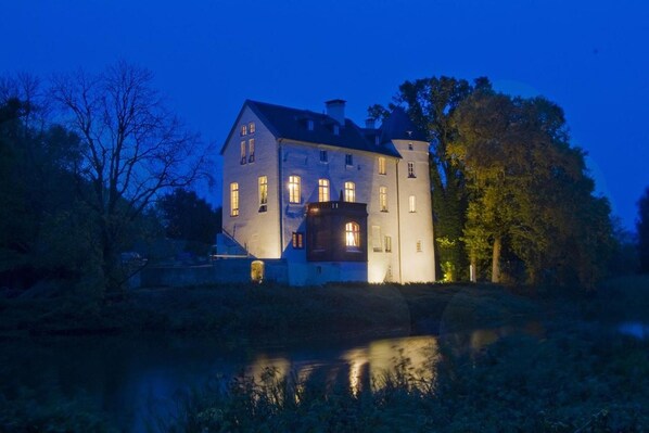 A large, illuminated building with multiple windows, situated by a body of water, surrounded by trees.