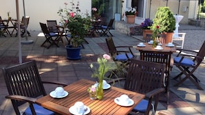 An outdoor patio with wooden tables and chairs, potted plants, and a small table with cups and saucers.