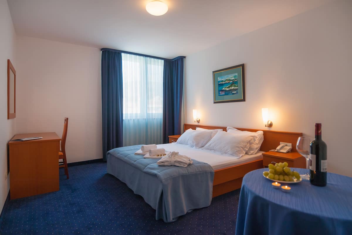 Habitación de hotel con una cama bien hecha, un escritorio con una silla, una mesita de noche con un teléfono y una botella de vino, y una pequeña mesa con un plato de uvas.