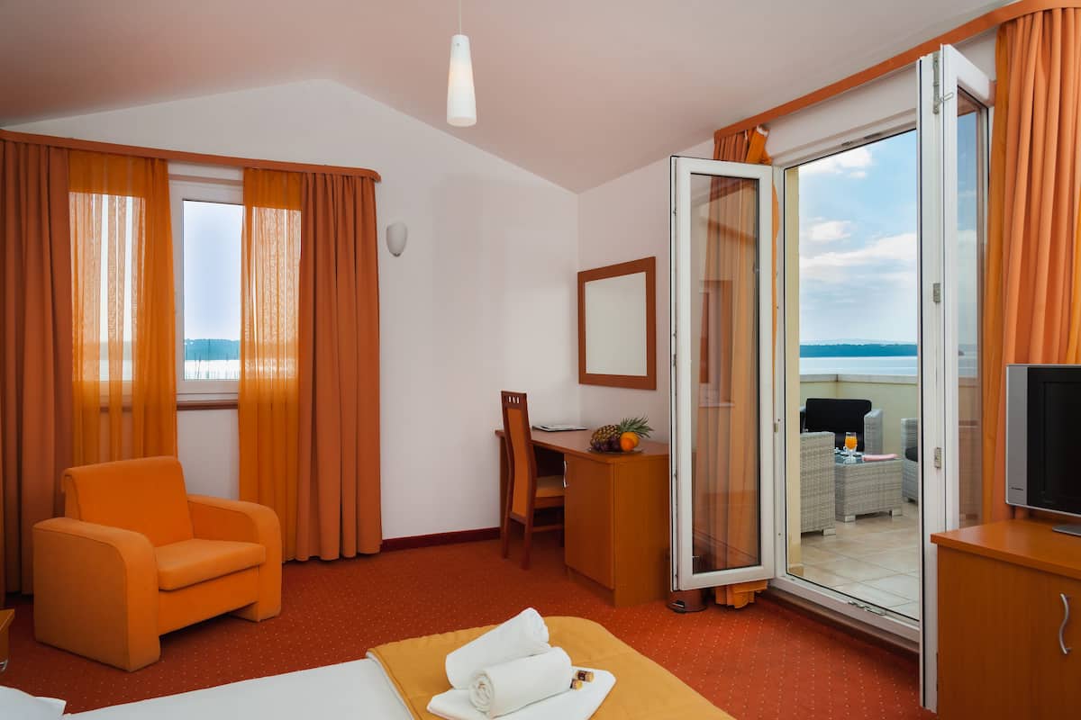 A hotel room with a bed, a desk, a chair, a television, and a sliding glass door leading to a balcony with a view of the sea.